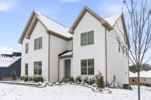 White brick two-story house with beige trim, large windows, and a snow-covered front yard under a blue winter sky.