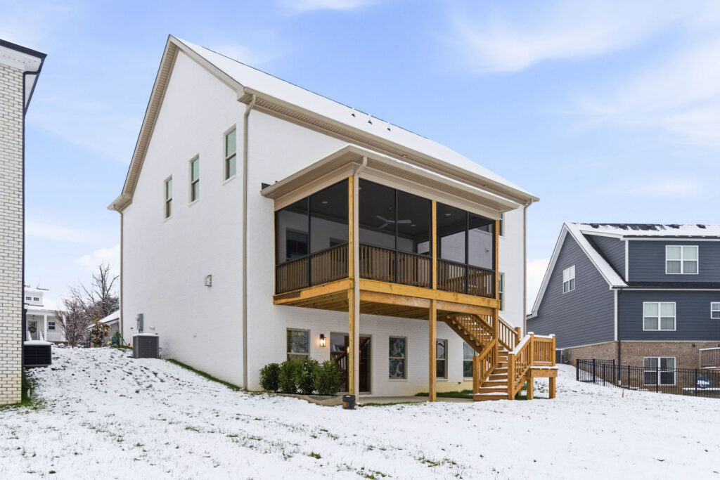 Backyard of a white brick house with a two-story wooden screened porch and staircase, snow on the lawn under a blue sky.