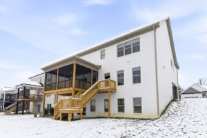White brick house with a large wooden deck, stairs, and screened porch, set in a snowy yard under a blue sky.