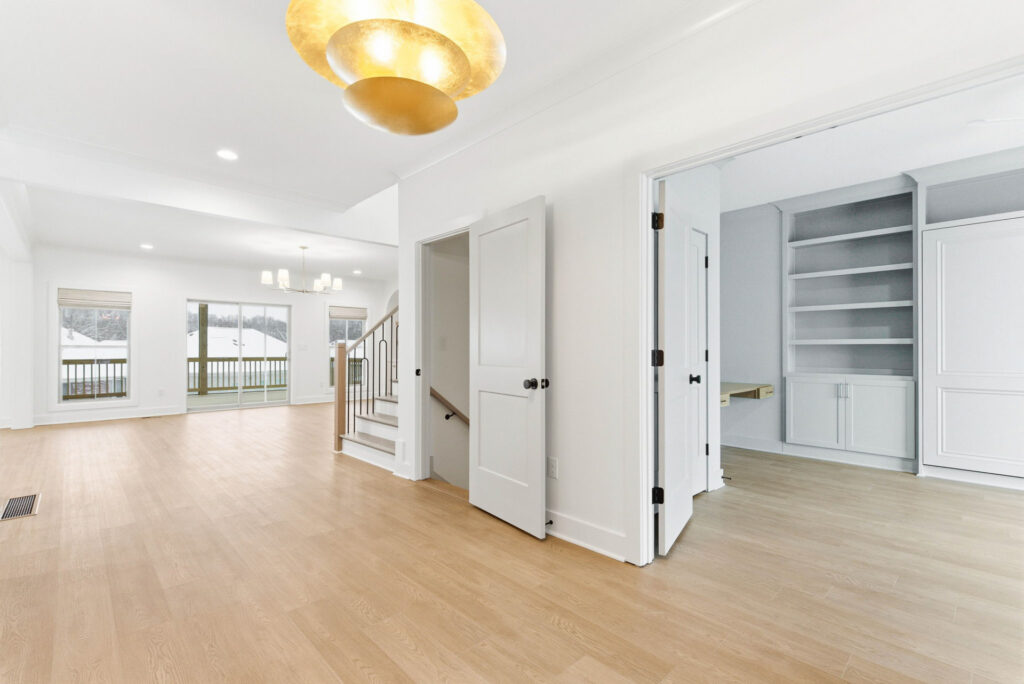 Bright open living area with light wood floors, white walls, and large windows leading to a balcony; staircase railing visible in the center.
