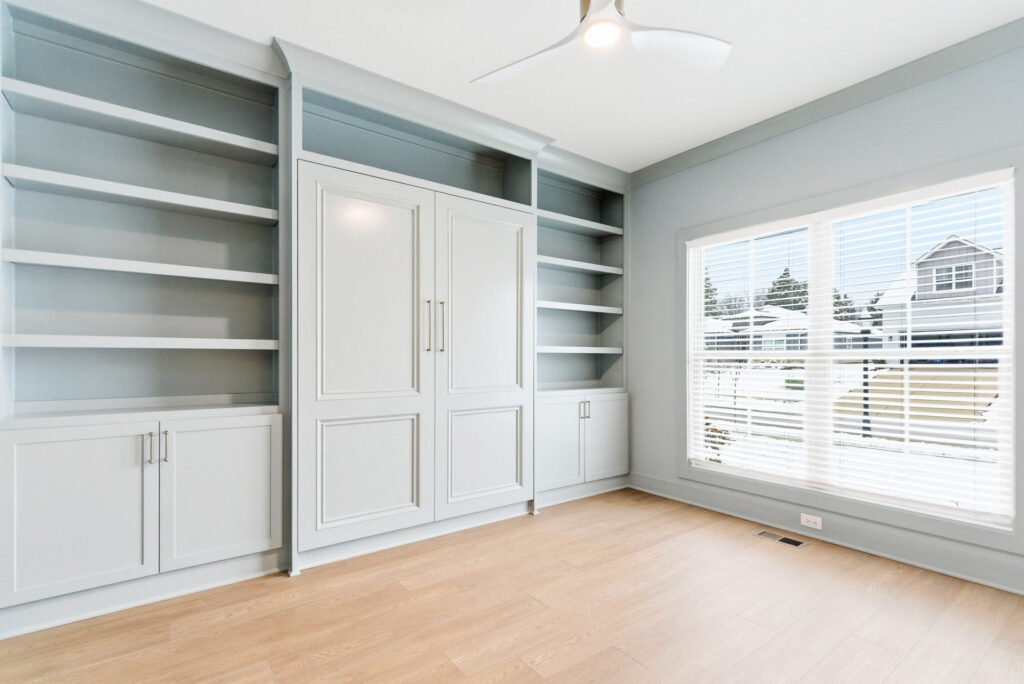 Bright laundry/storage room with gray built-in cabinets and open shelving beside a large window with blinds.